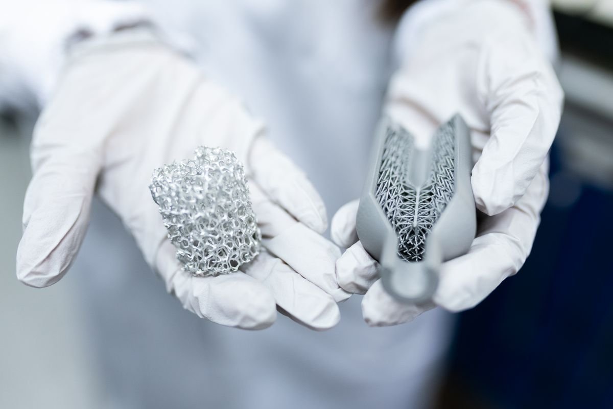 Gloved hands holding two 3D-printed metal parts with organic lattice and topology-optimized structures
