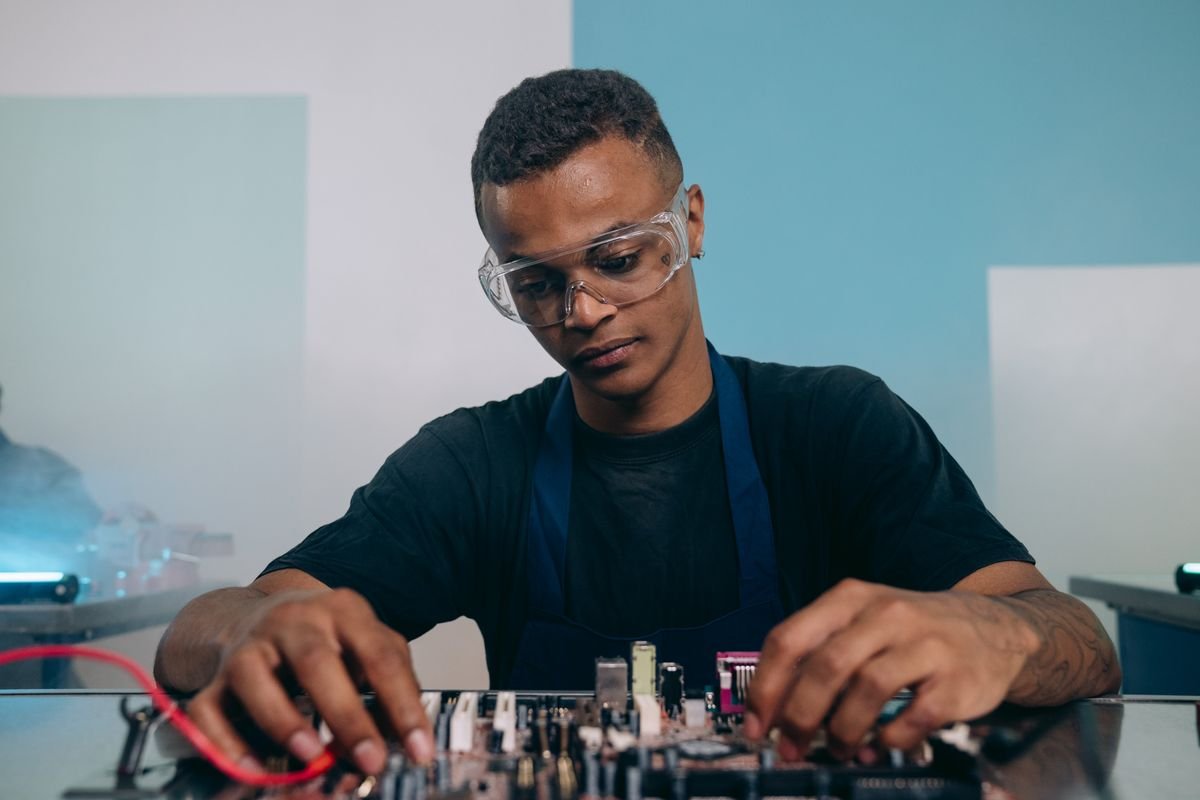 A technician in safety glasses working on a circuit board at an electronics bench