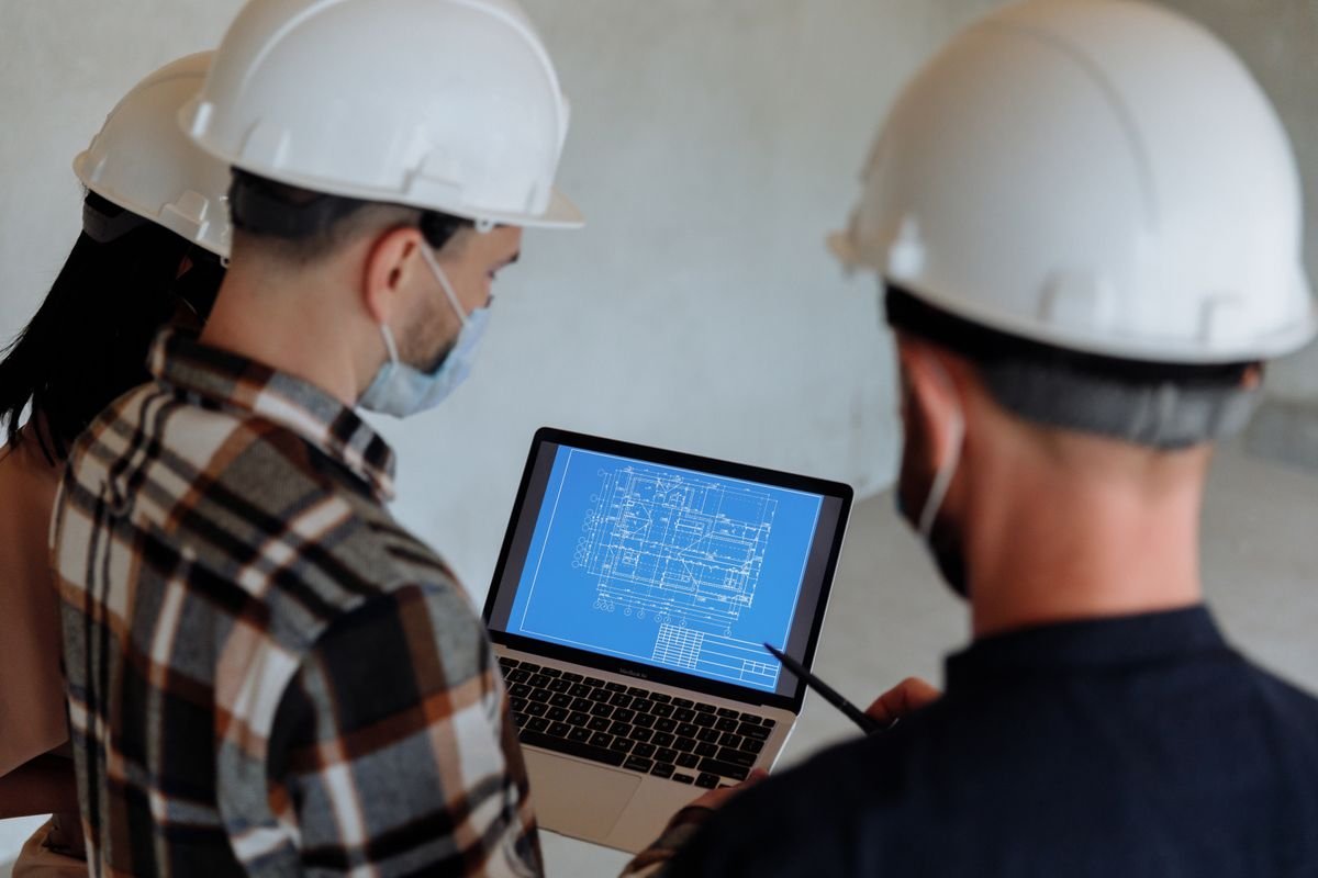 Two engineers in hard hats reviewing a construction blueprint on a laptop
