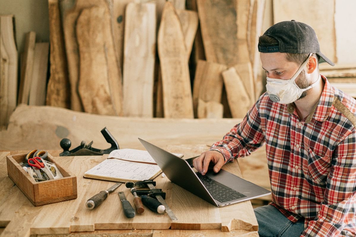 A maker in a workshop using a laptop beside hand tools and timber